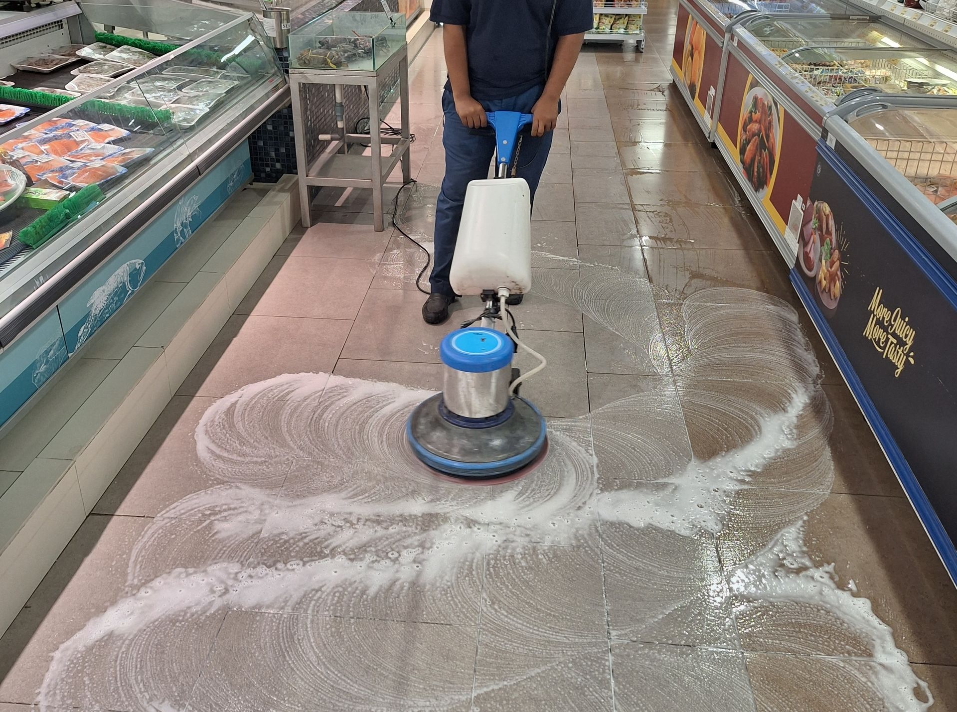 Worker cleaning a store aisle floor with a floor buffer, leaving wet swirls on the tile.