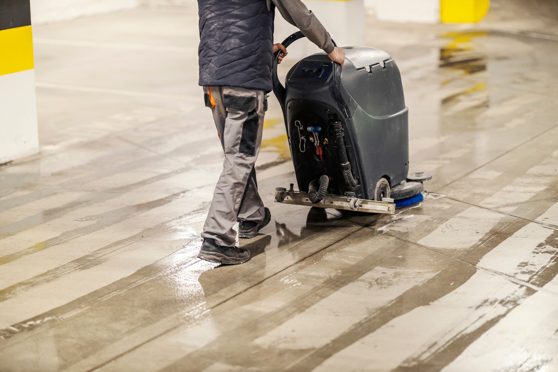 Person pushing a gray floor-cleaning machine across a wet indoor floor.
