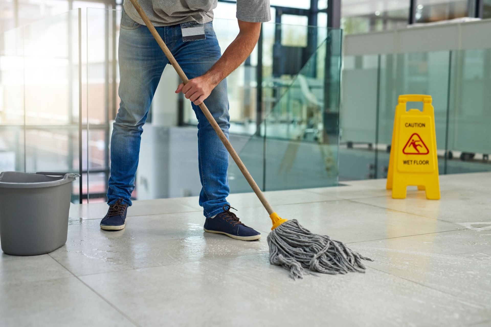 Person mopping a glass lobby floor beside a yellow wet floor sign and bucket