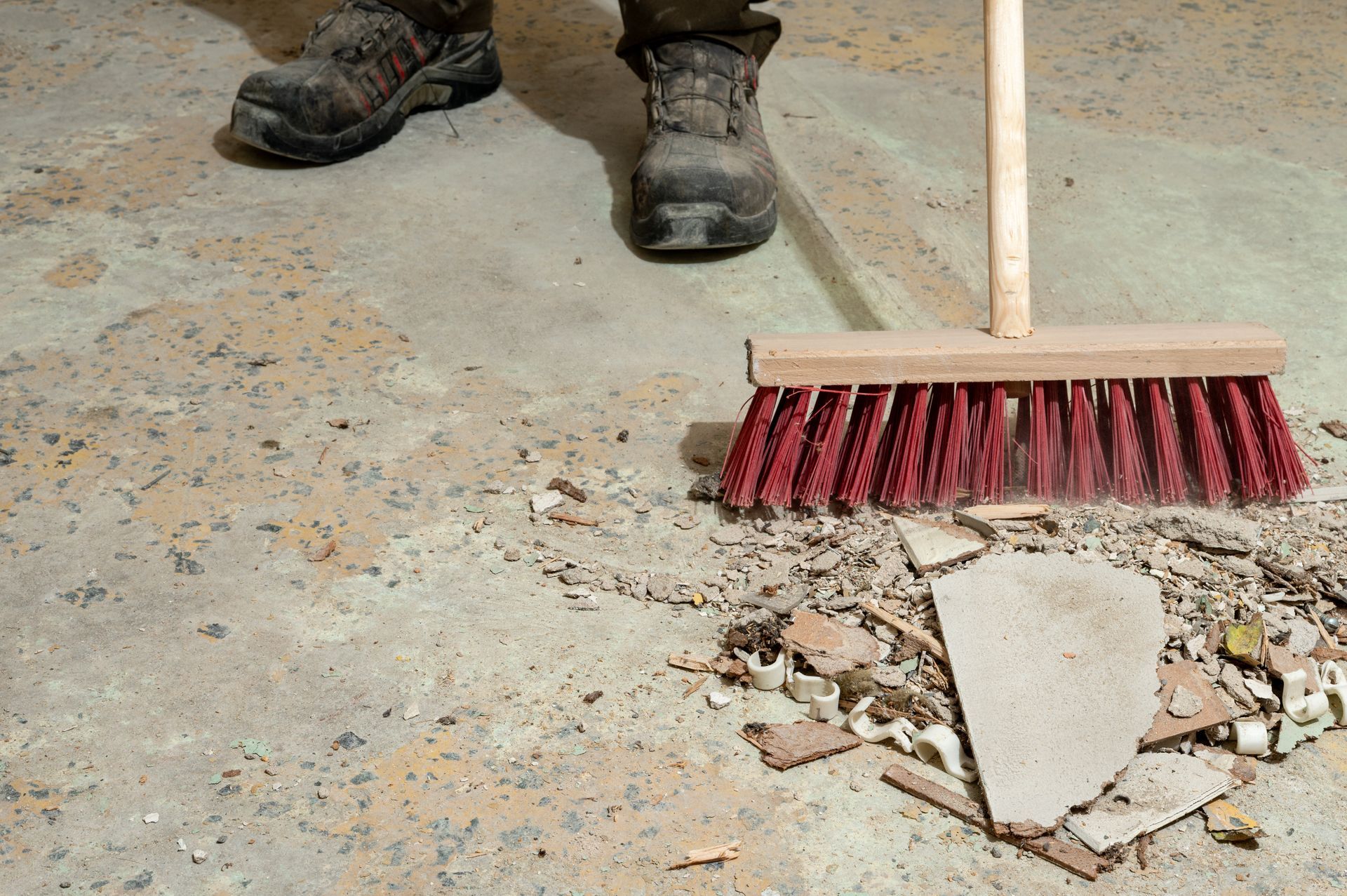 Red broom sweeping broken concrete and dust near work boots on a dusty floor
