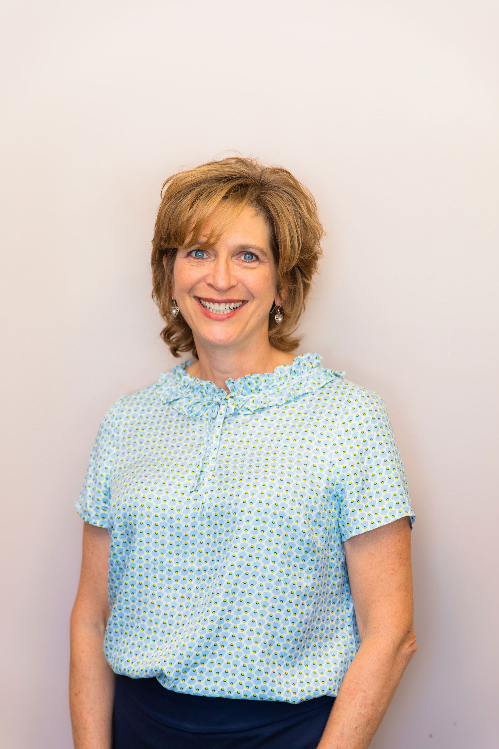 A woman in a blue shirt is smiling and standing in front of a white wall.