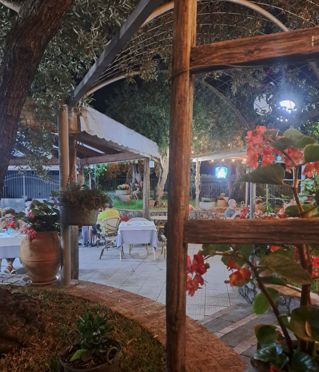 Caffè all'aperto serale con sedie bianche, luci calde e fiori rossi sotto un arco di legno.