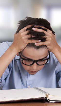 Un jeune garçon portant des lunettes est assis à un bureau avec ses mains sur la tête.
