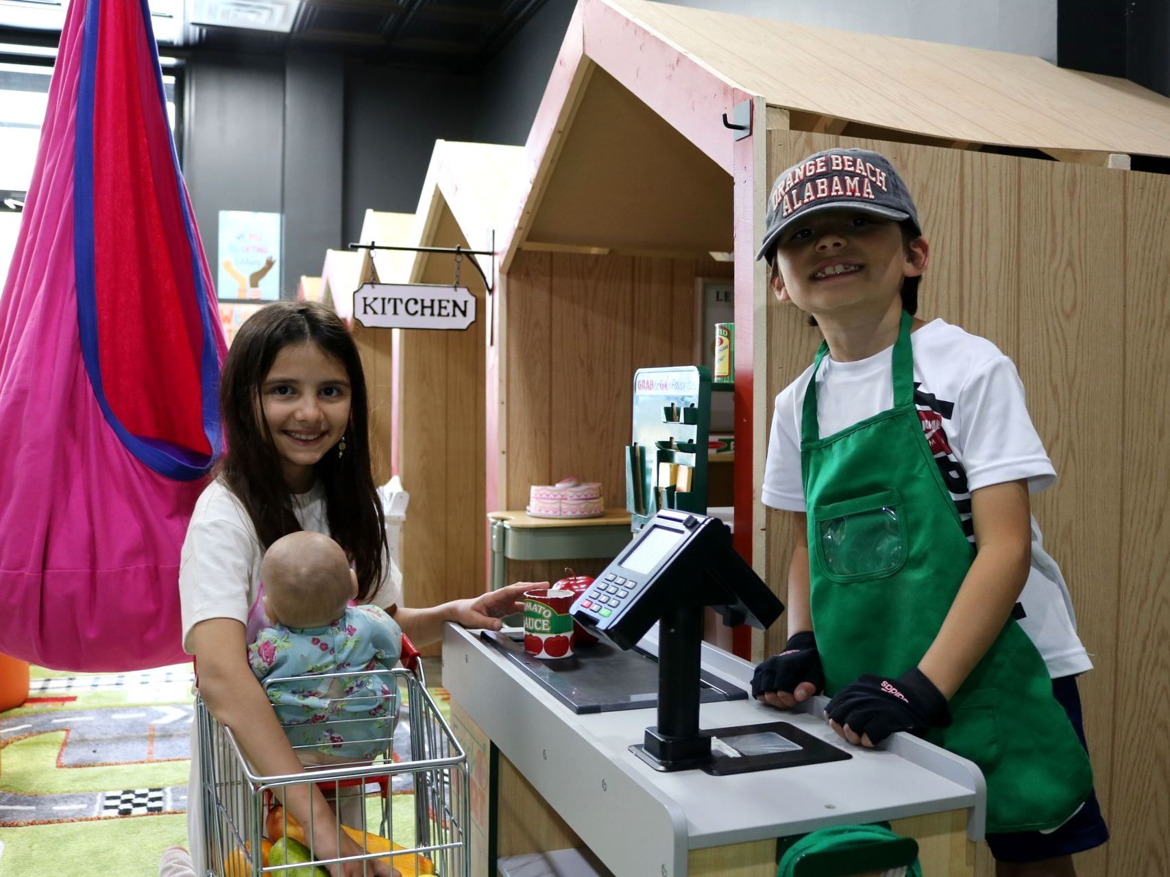 A girl is holding a baby and a boy is standing in front of a cash register.
