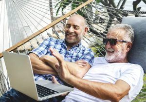 Two men are laying in a hammock looking at a laptop.