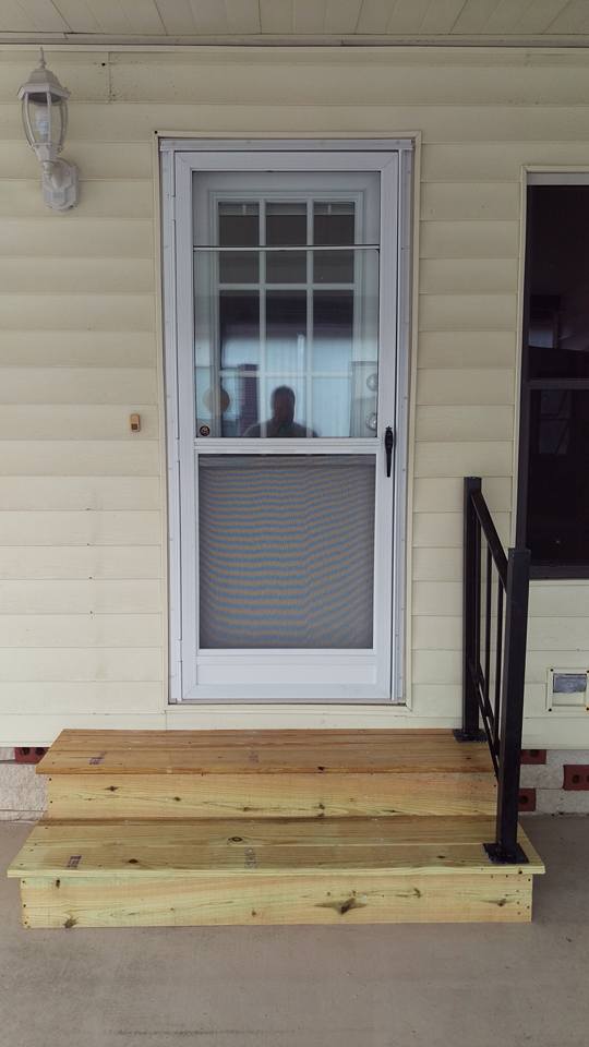 Wooden steps lead to a screened white door on a beige house. A black railing is on the right.
