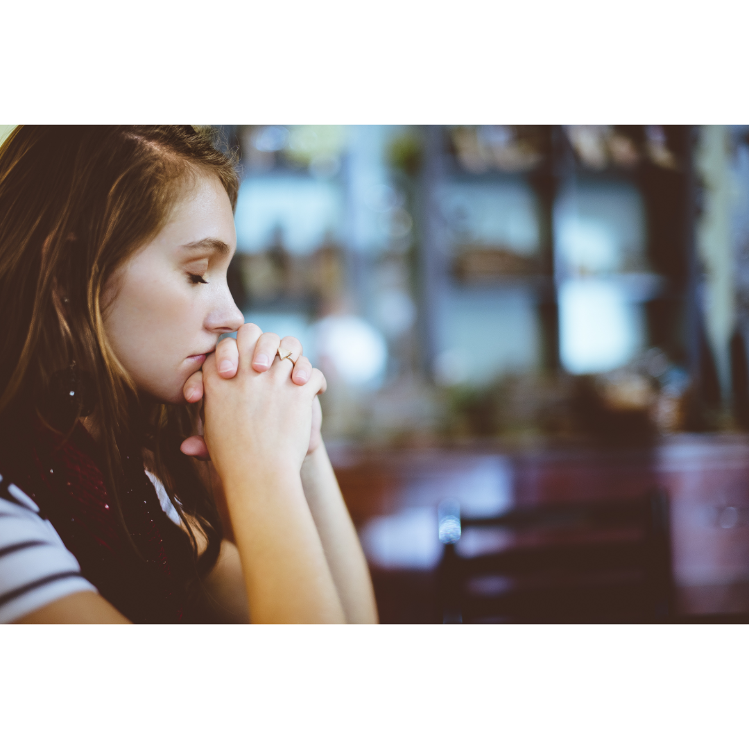 A woman is sitting at a table with her hands folded in prayer.