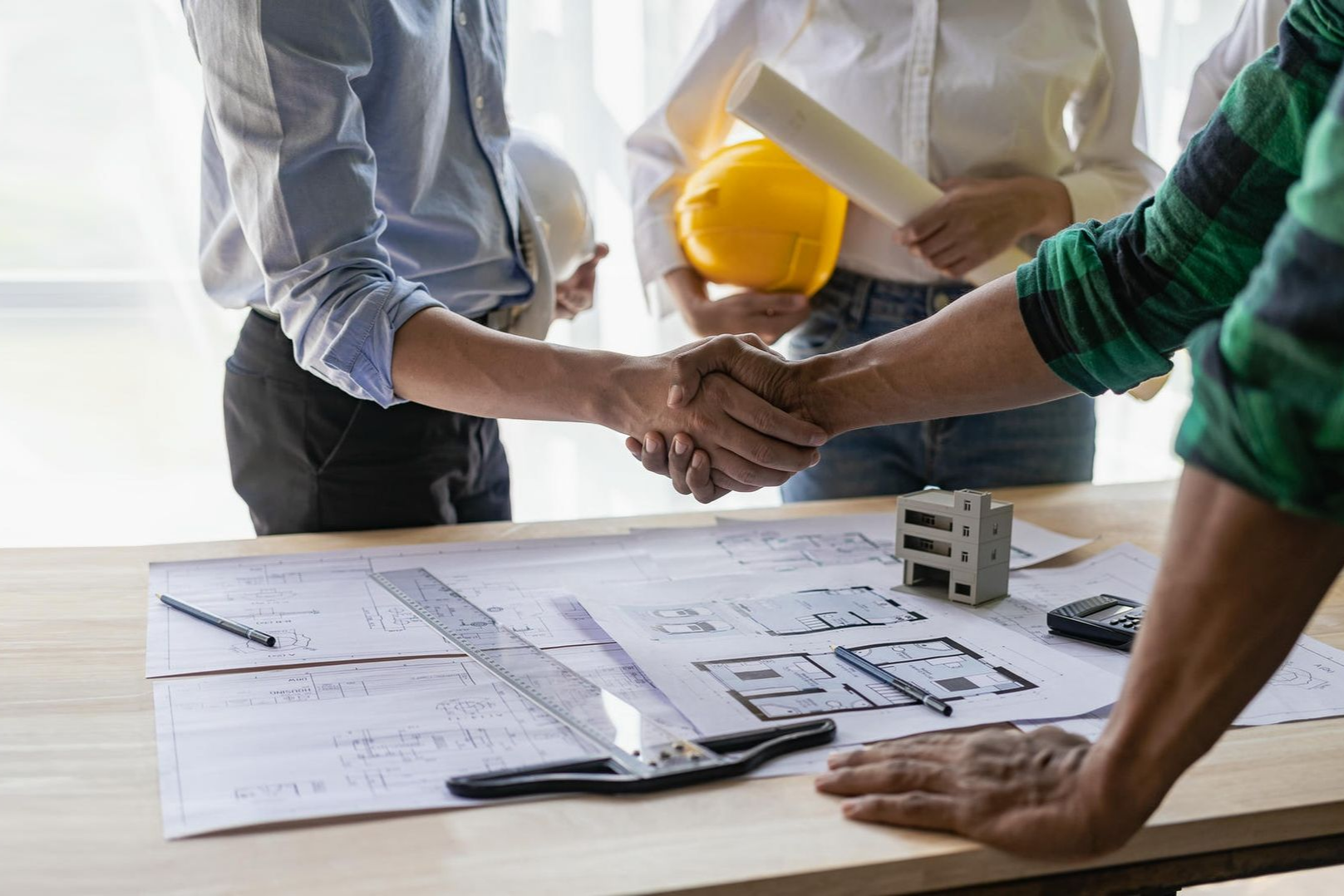 People shaking hands over blueprints at a desk with construction tools.