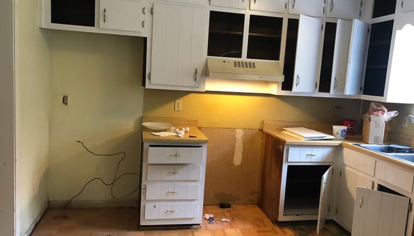 Kitchen undergoing renovation; white cabinets, bare walls, exposed wiring, and cabinetry.