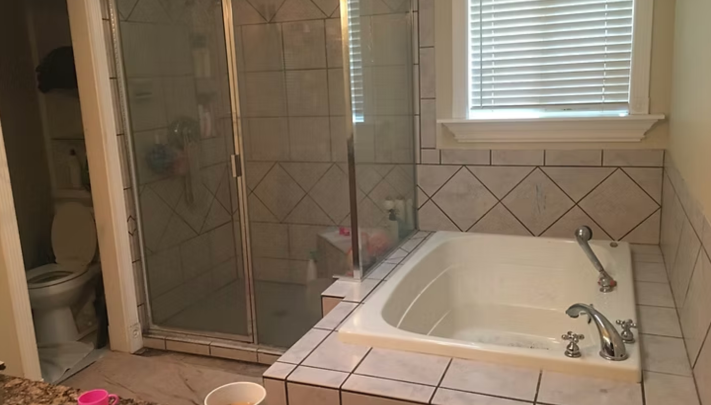Bathroom with white bathtub, glass shower, and tiled walls. Window with blinds. Toilet on left.