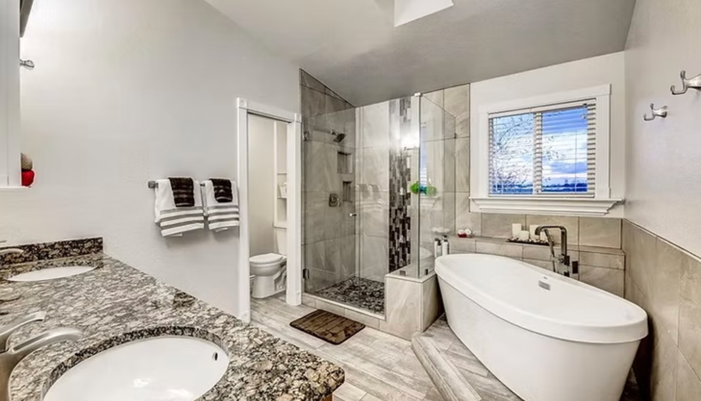 Modern bathroom with a bathtub, shower, double vanity, and a window overlooking a view.