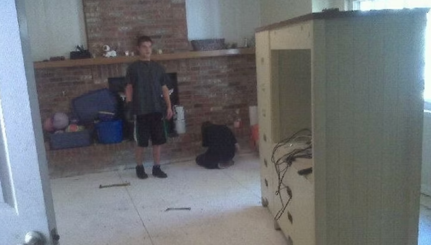 Person standing in a room with a fireplace, a cabinet, and another person on the floor.