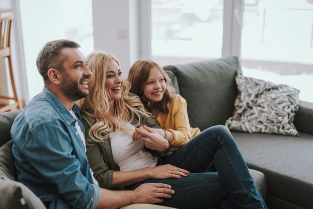 Three people sit smiling on a gray couch in a bright living room.
