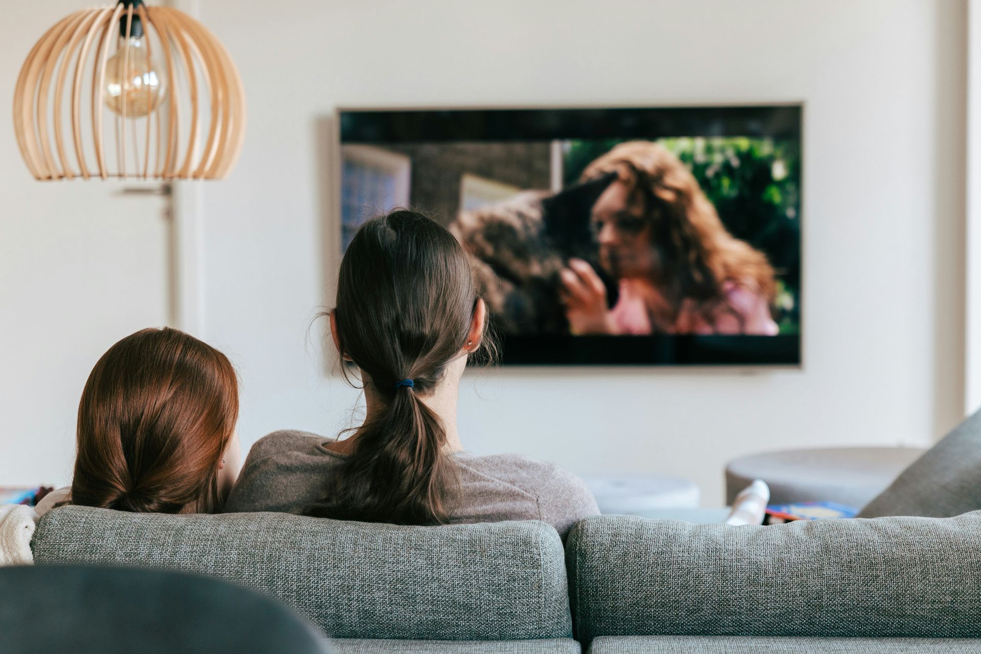 Two people sit on a gray couch watching TV in a bright living room.