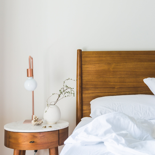 Nightstand with lamp, vase of greenery, and a bed with white sheets.