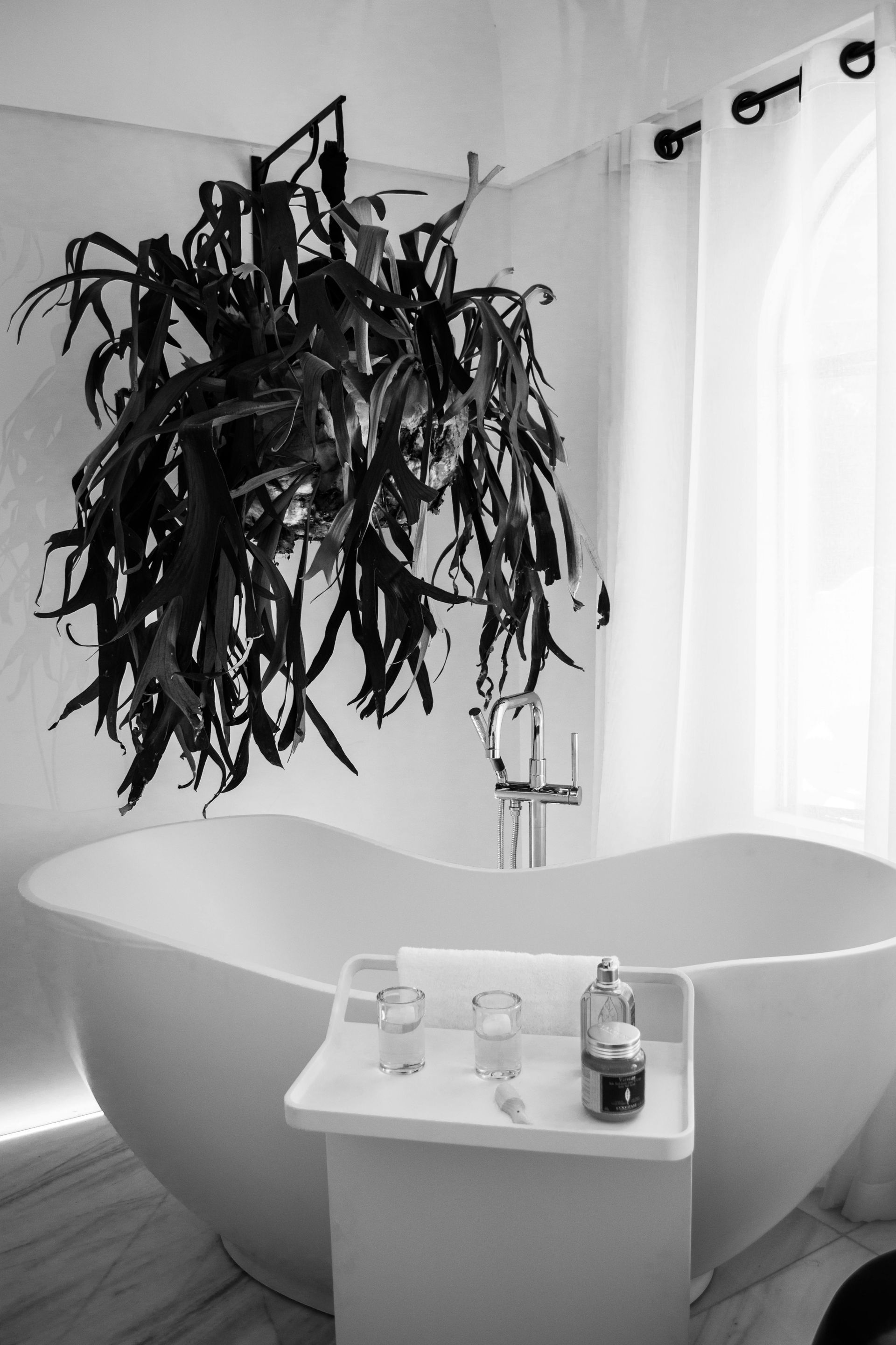 Freestanding white bathtub with hanging plant, side table with spa products, and arched window.