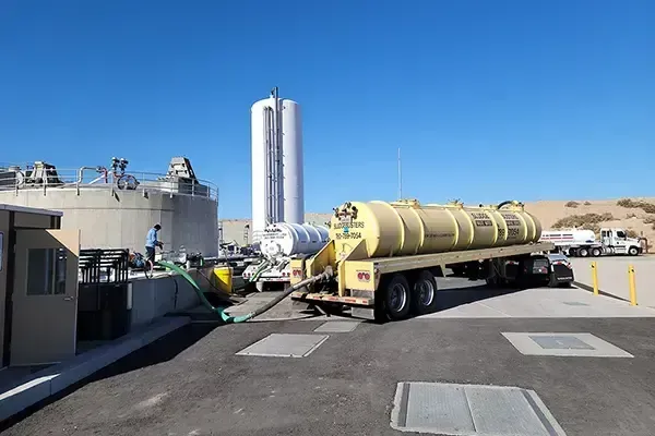 A tanker truck pumps liquid waste at a treatment plant with tall white silo, tan tanks, and a clear blue sky.