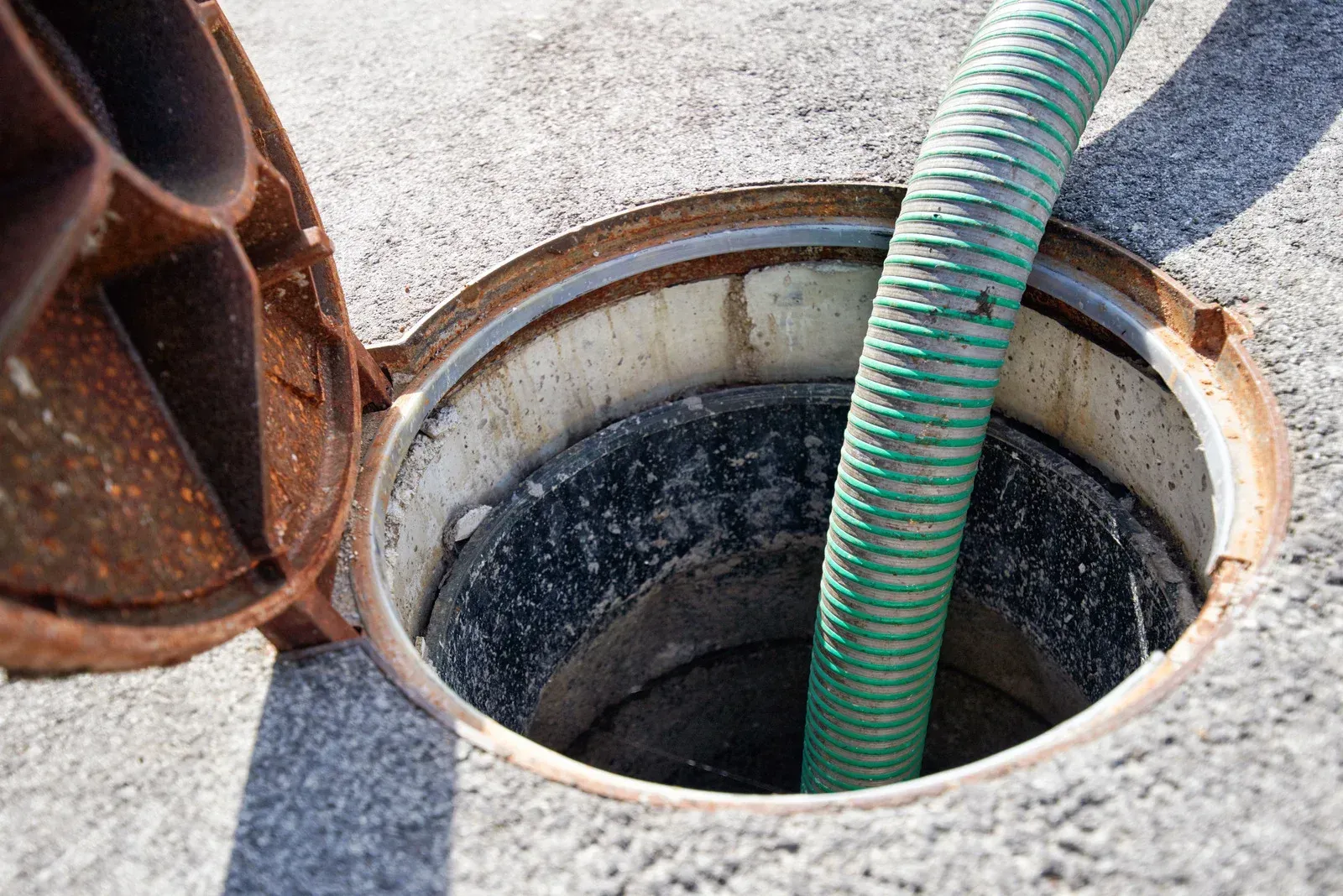 Open manhole with a green hose inside; lid is ajar. Concrete, outdoor setting.