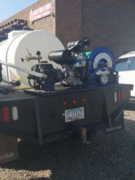 Water truck with tank, pump, and hose reel on a flatbed. Outdoors, sunny.
