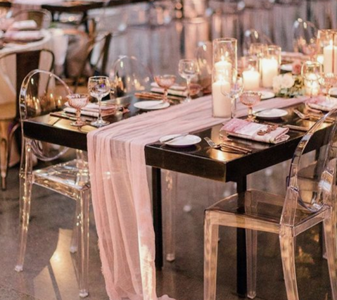 A table set for a wedding reception with a pink table runner and clear chairs.