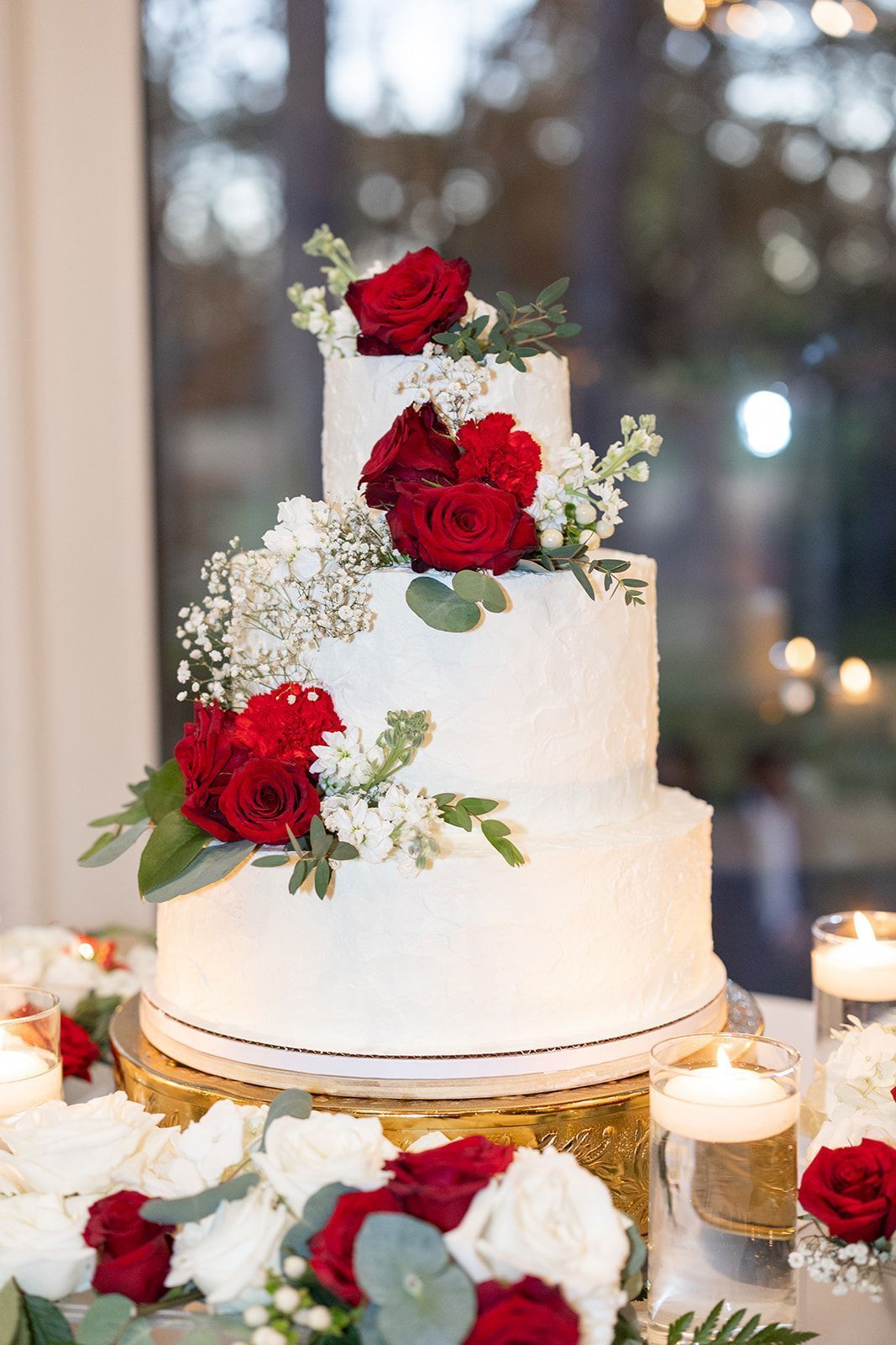 A white wedding cake with red and white flowers and candles on a table.