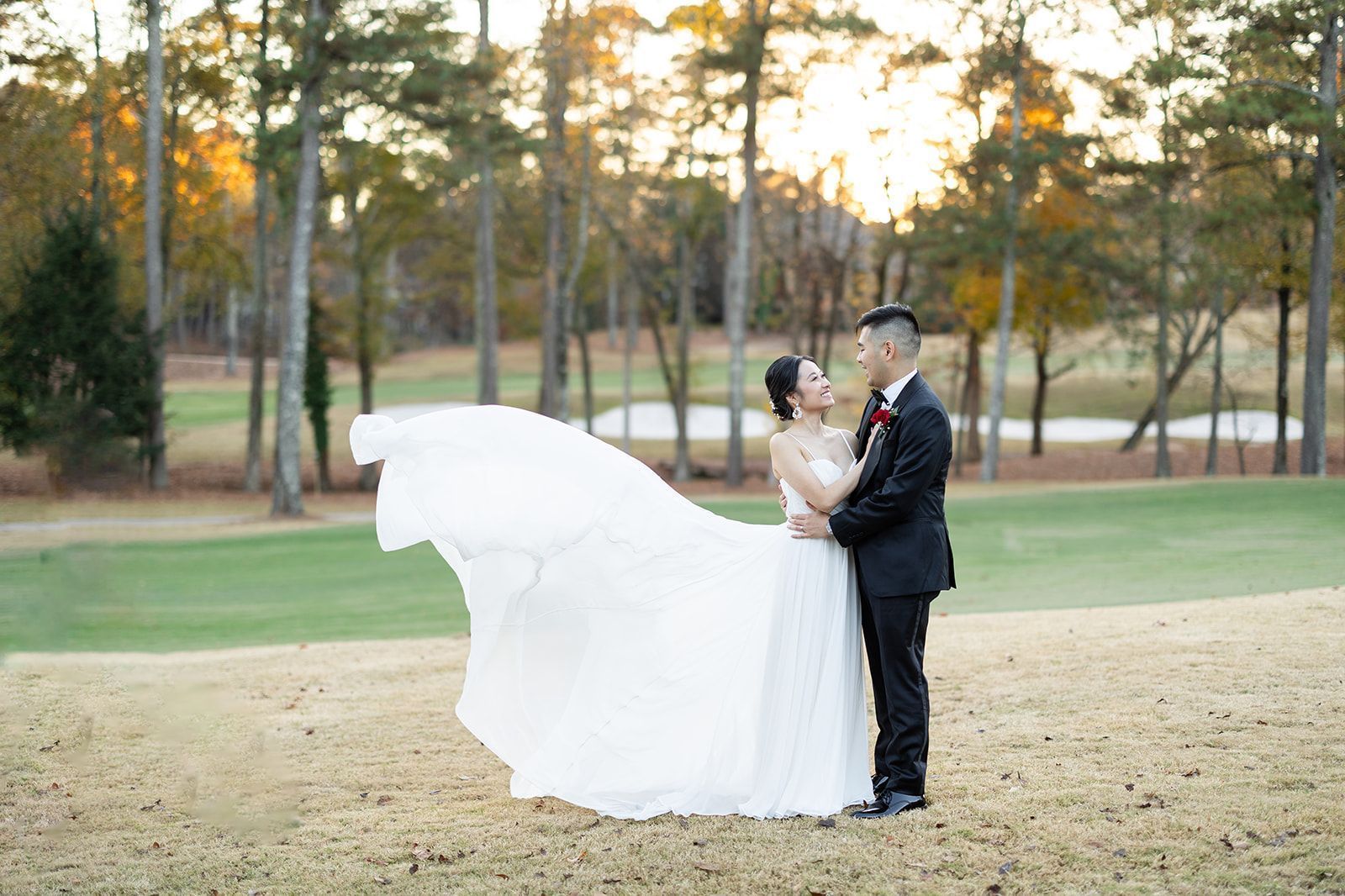 A bride and groom are standing in a field with their wedding dress blowing in the wind.