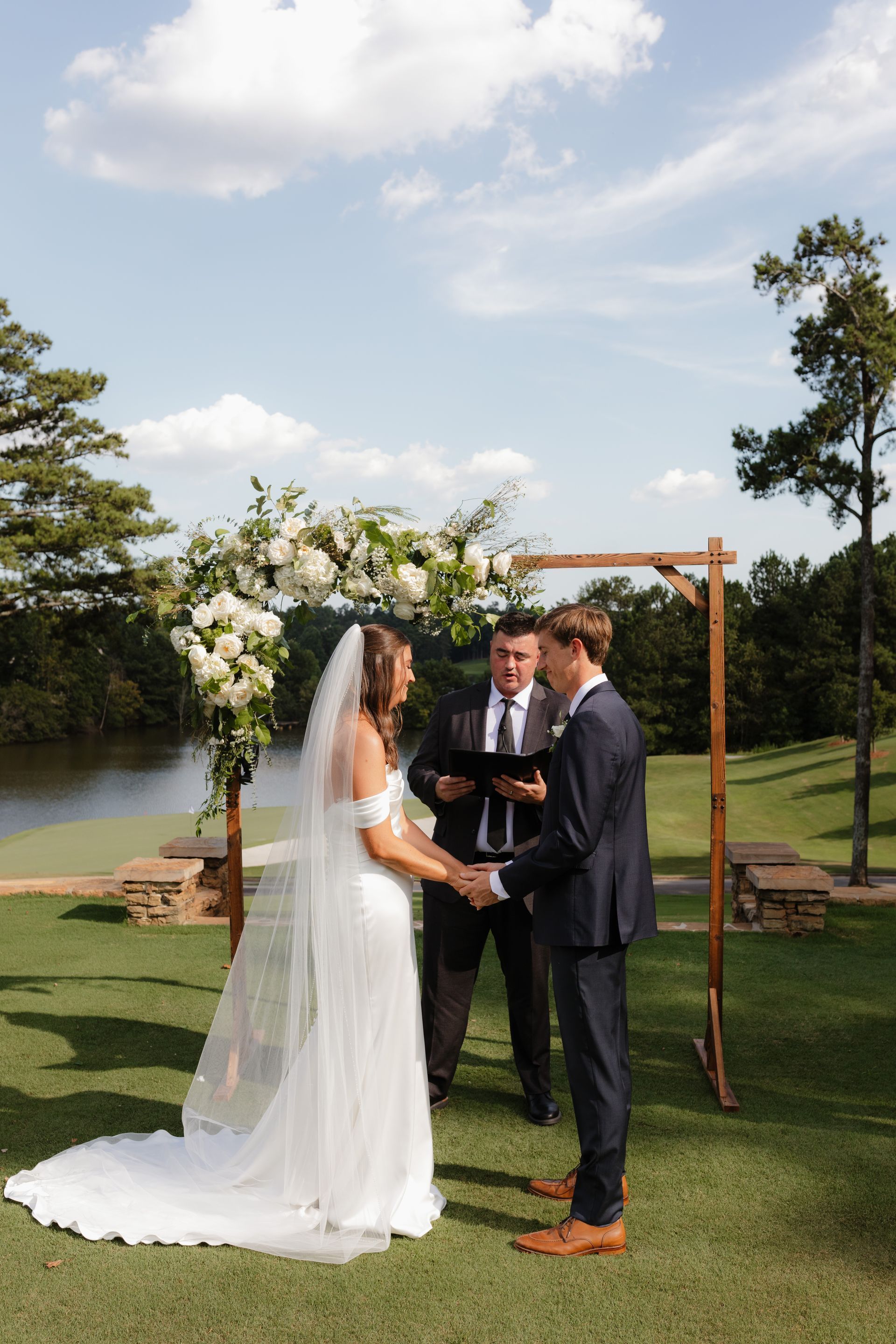 A bride and groom are holding hands during their wedding ceremony in front of a lake.