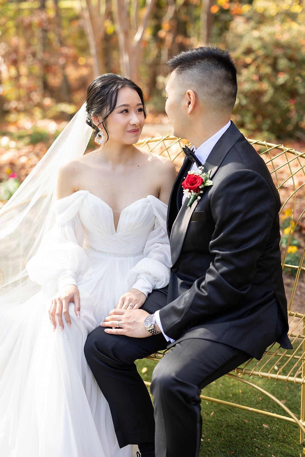 A bride and groom are sitting on a bench holding hands and looking at each other.