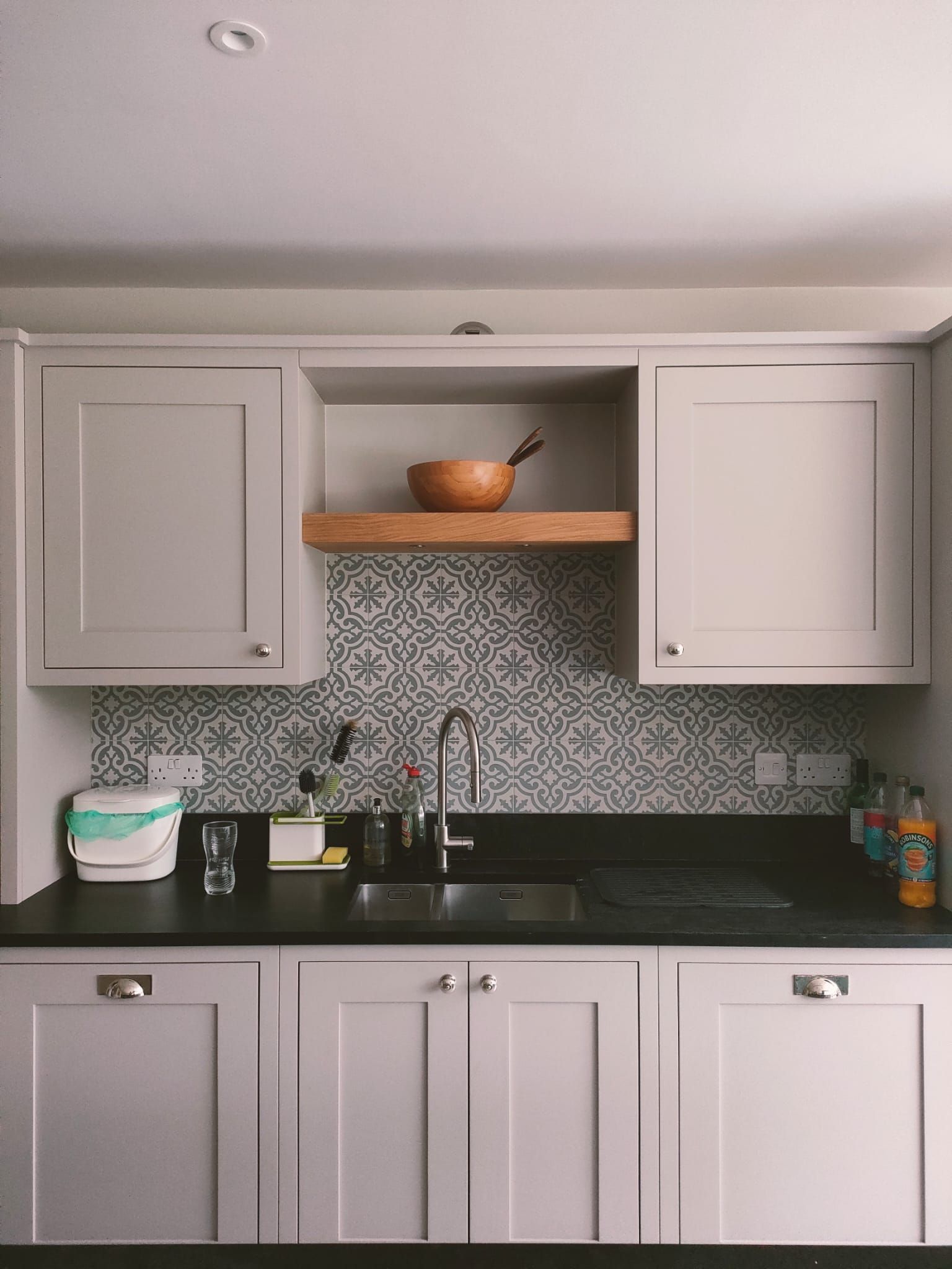 A kitchen sink area with light grey cabinets, a patterned tile backsplash, a wooden shelf, and a dark countertop.