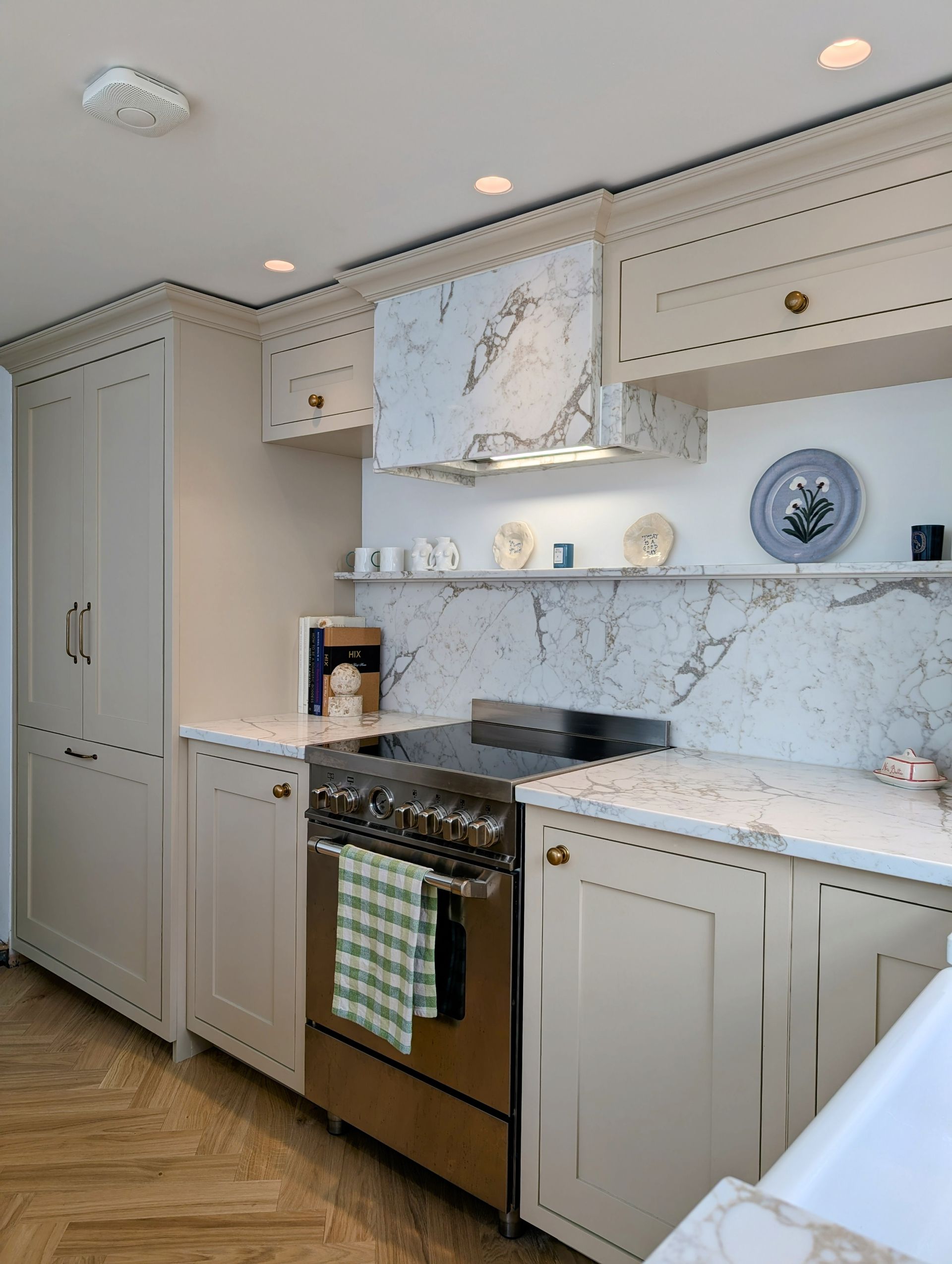 A neutral-toned kitchen featuring a marble backsplash, matching range hood, stainless steel stove, and herringbone floor.