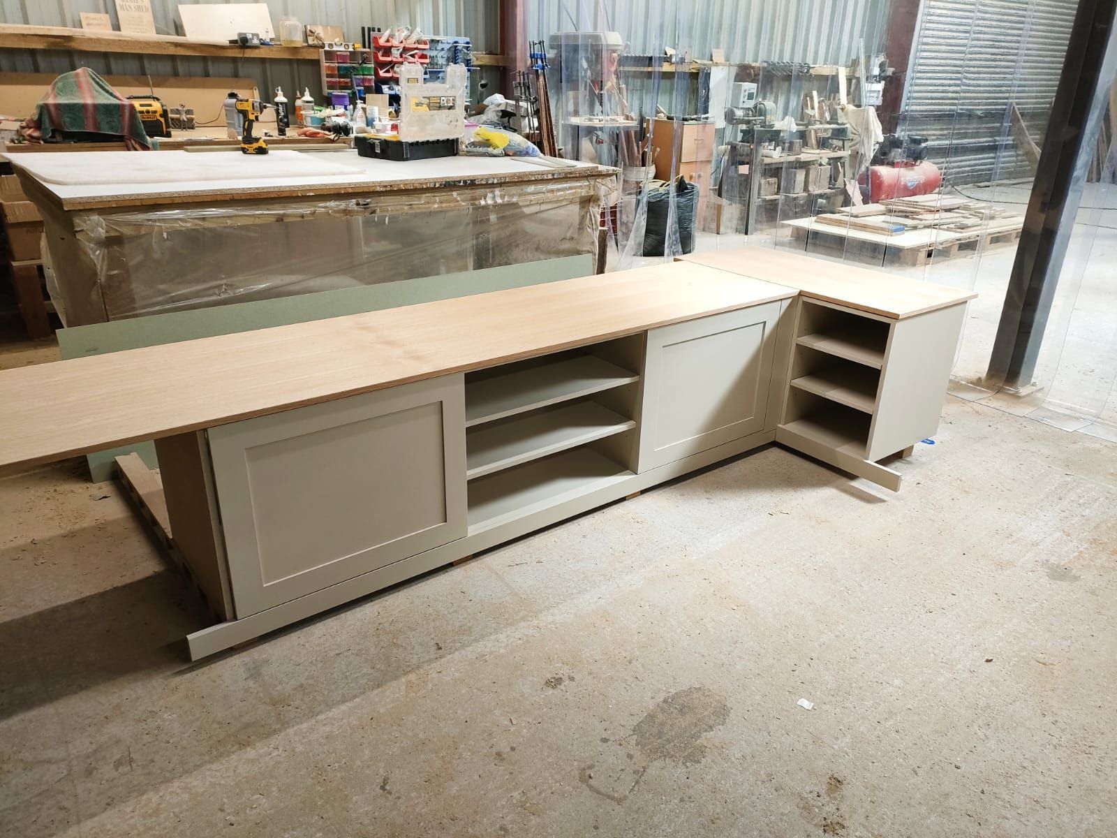 A partially assembled beige wooden cabinet with shelving sits in a workshop, topped with a piece of plywood.