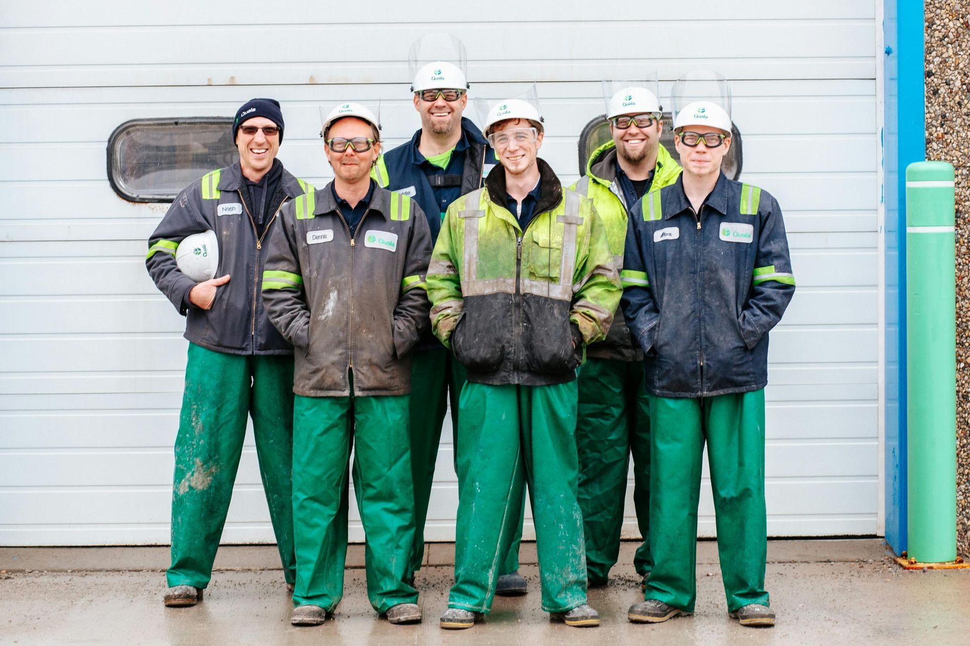 Six people wearing hard hats, matching work jackets, and green pants stand in a line before a white industrial door.