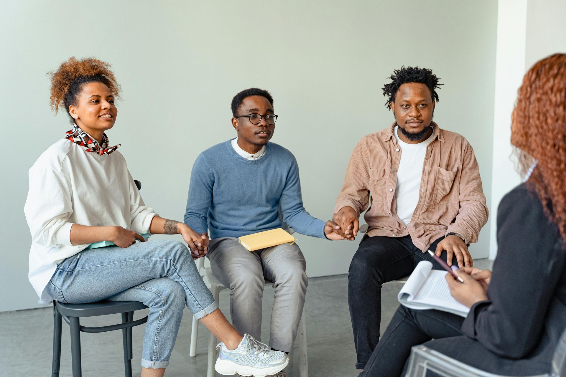 A group of people sit in a circle holding hands while talking in a simple, well-lit room.
