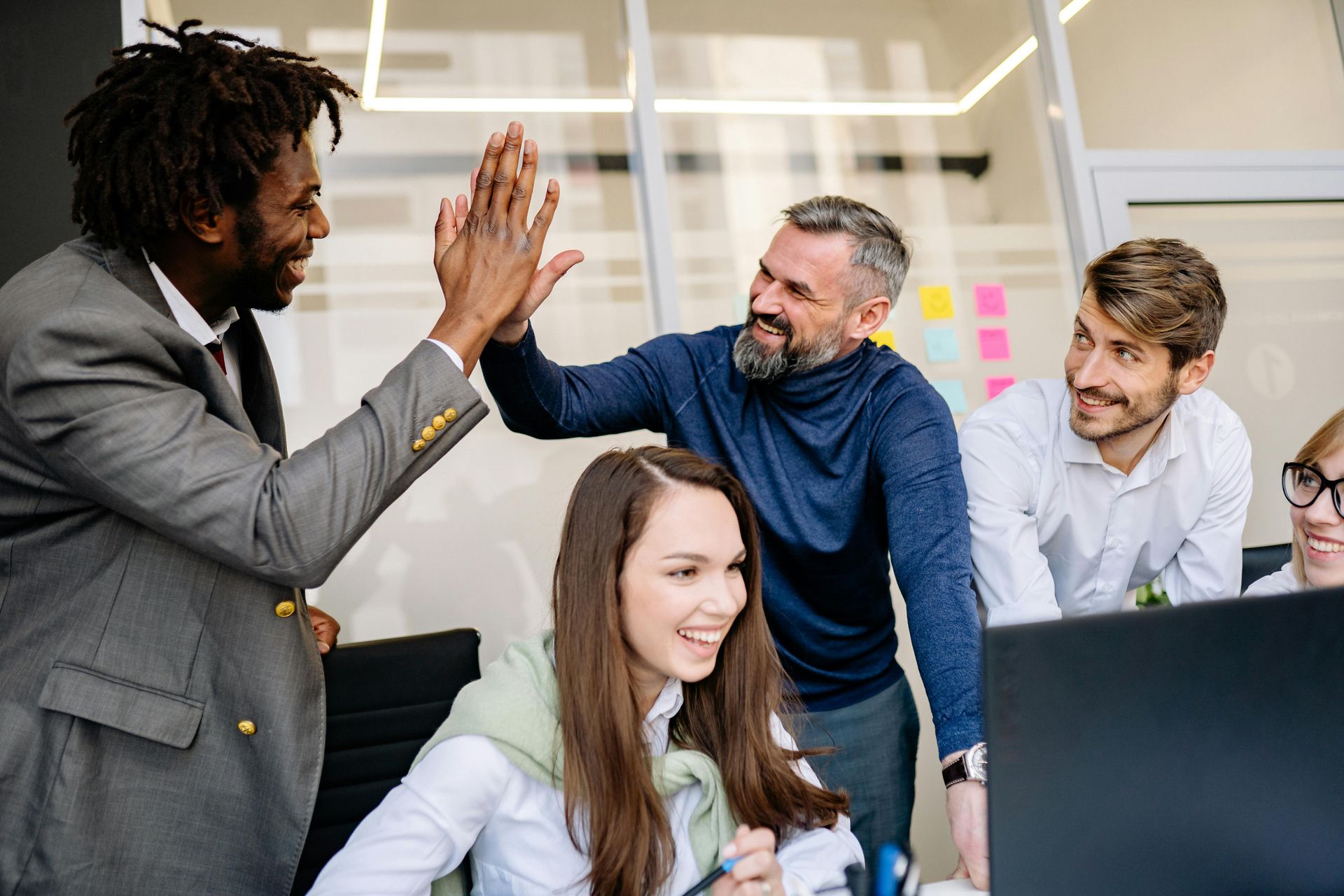 A diverse professional team in an office high-fiving and smiling while gathered around a computer screen.