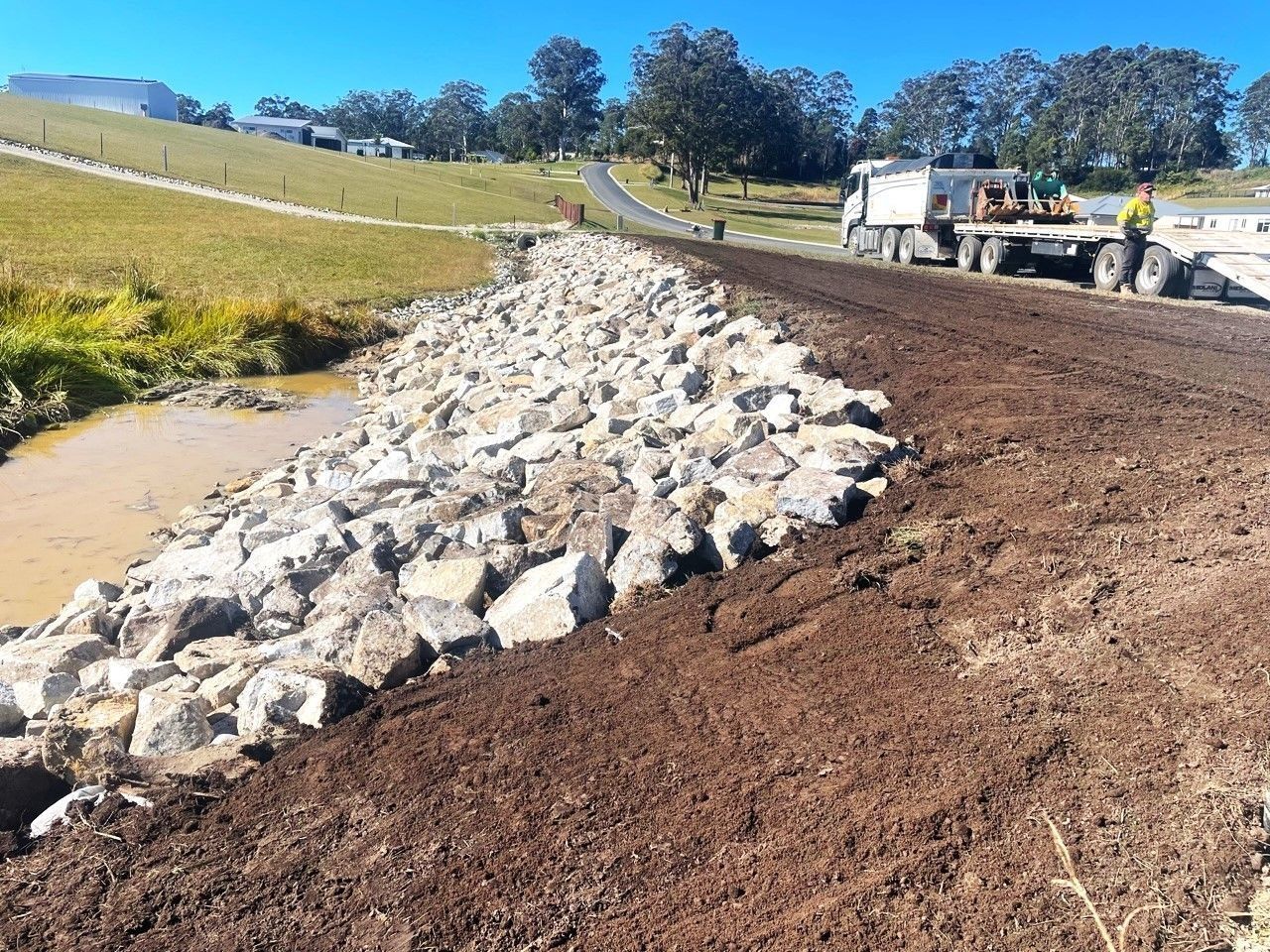 Rocks Lining a Stream Next to a Dirt Road, With a Truck — Blue Dog Earthmoving Landscape Supplies in Urunga, NSW