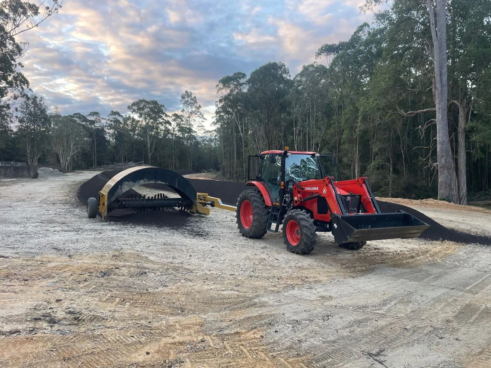 Red Tractor Pulling a Black Compost Spreader on a Gravel Road — Blue Dog Earthmoving Landscape Supplies in Bowraville, NSW