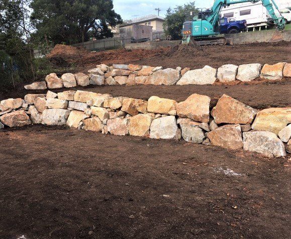 Stone Retaining Walls on a Hillside With an Excavator in the Background — Blue Dog Earthmoving Landscape Supplies in Scotts Head, NSW