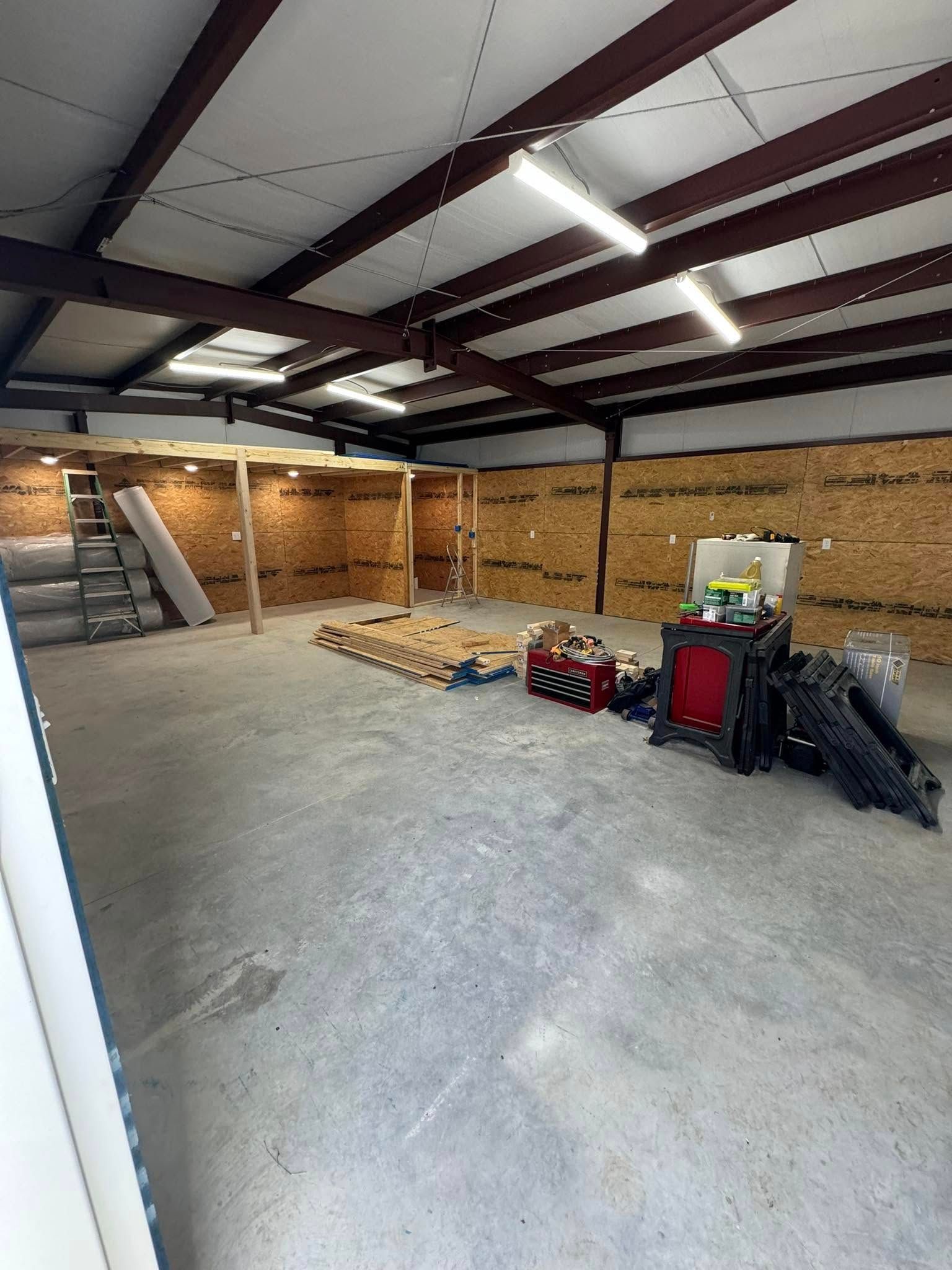 Interior view of a metal building under construction. Walls are partially covered in wood. Tools and materials are scattered around the concrete floor.