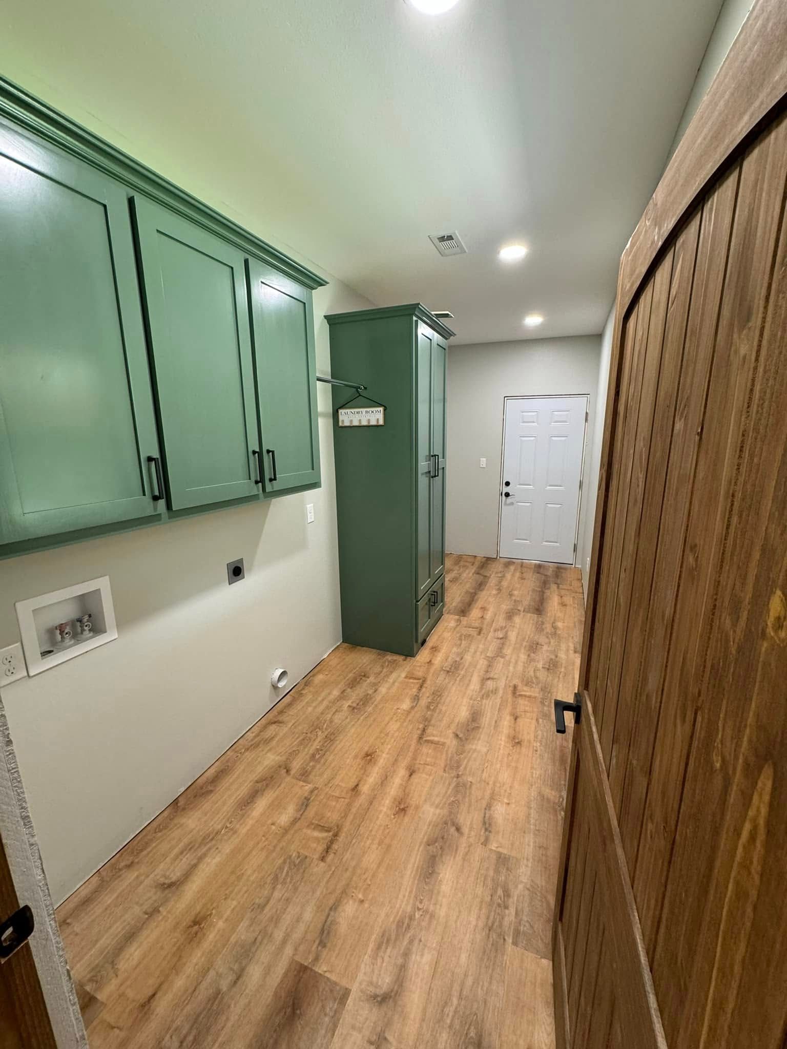 A laundry room with green cabinets and a wooden door, featuring wood-look flooring. The space has a tall storage cabinet and a white door.