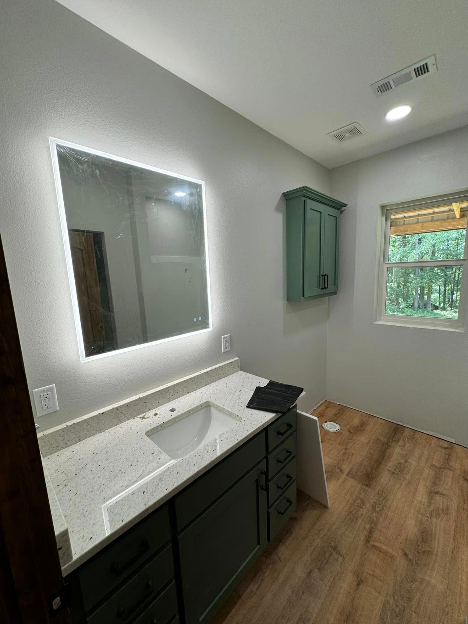 Modern bathroom with a lighted mirror, green cabinets, and a window overlooking trees.