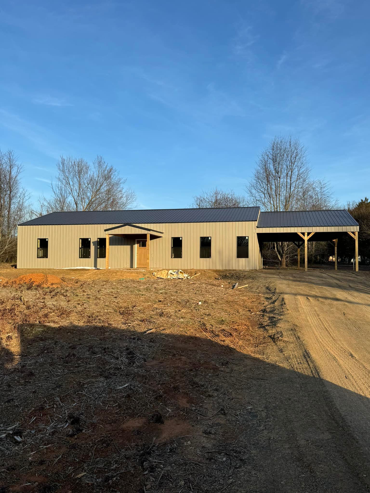 A light-colored, wooden building with a dark roof, possibly a barn, on a dirt lot with a clear blue sky.