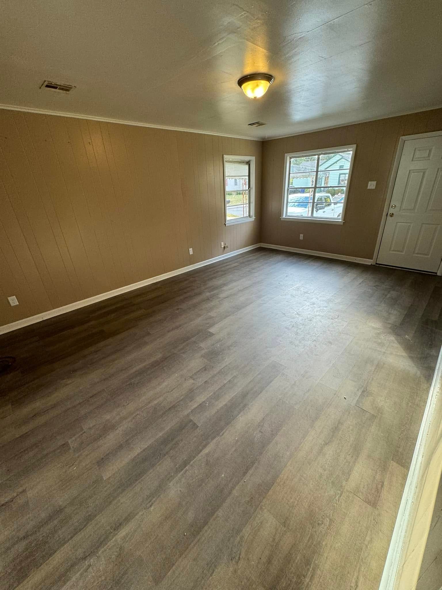 Empty living room with brown walls, wood-look flooring, two windows, and a white door.