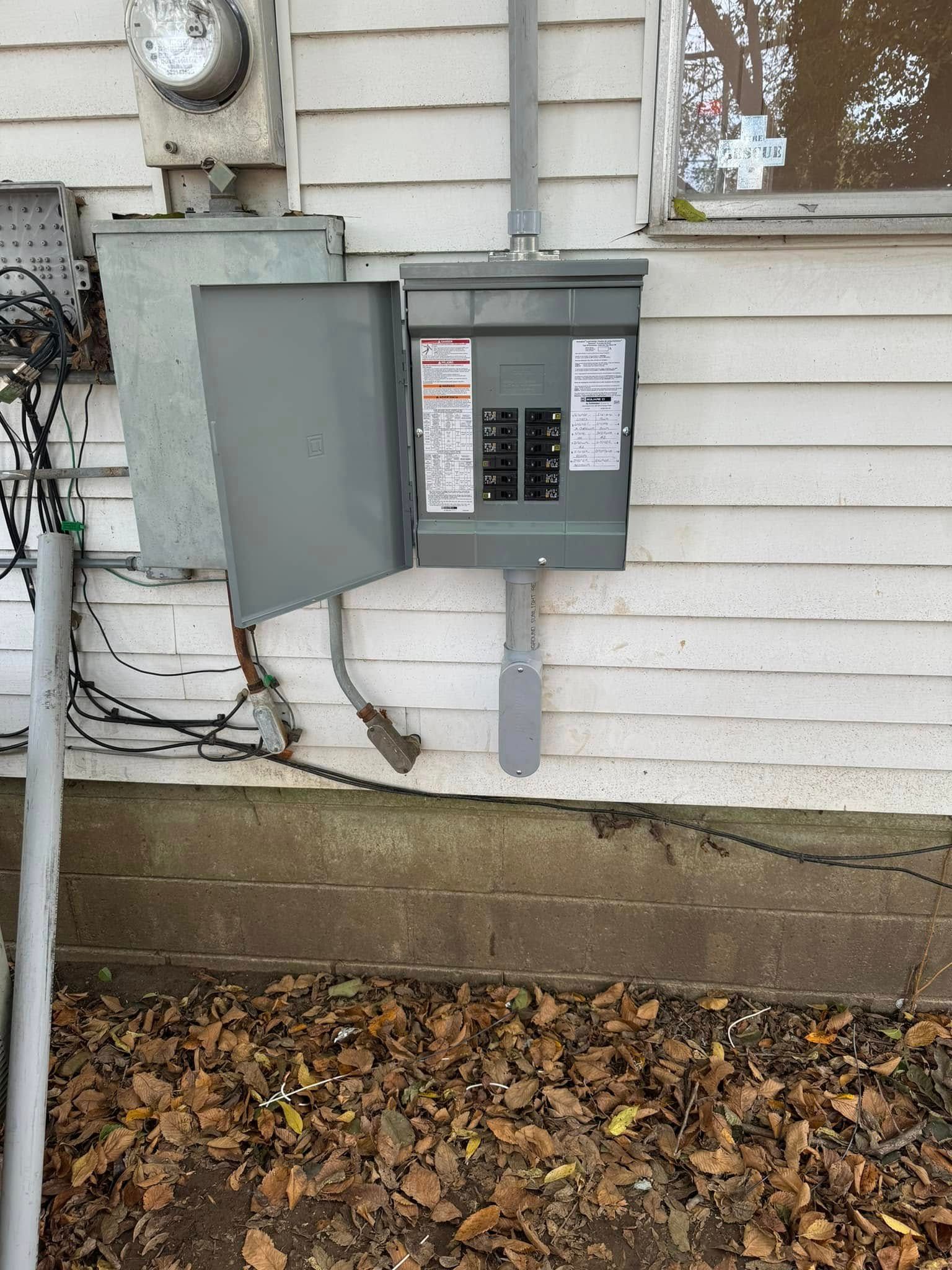 Electrical panel on the exterior of a building with its door open, revealing circuit breakers. Grey panel and siding.