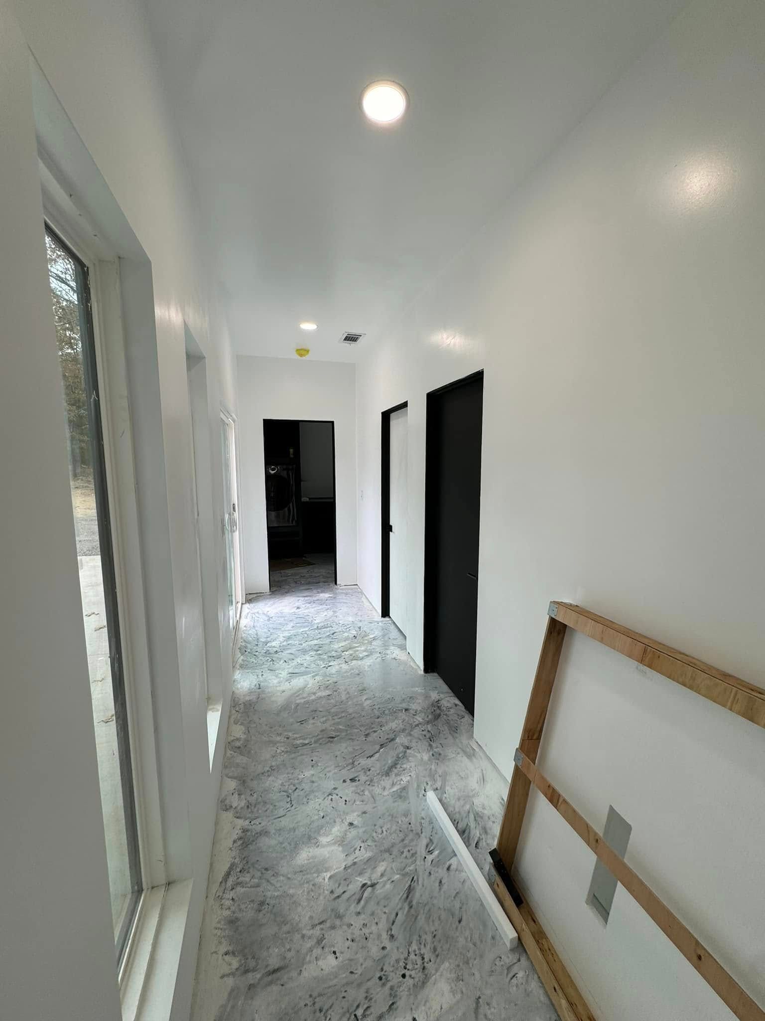 Narrow white hallway with light gray speckled flooring, black doors, and an unfinished artwork leaning against the wall.