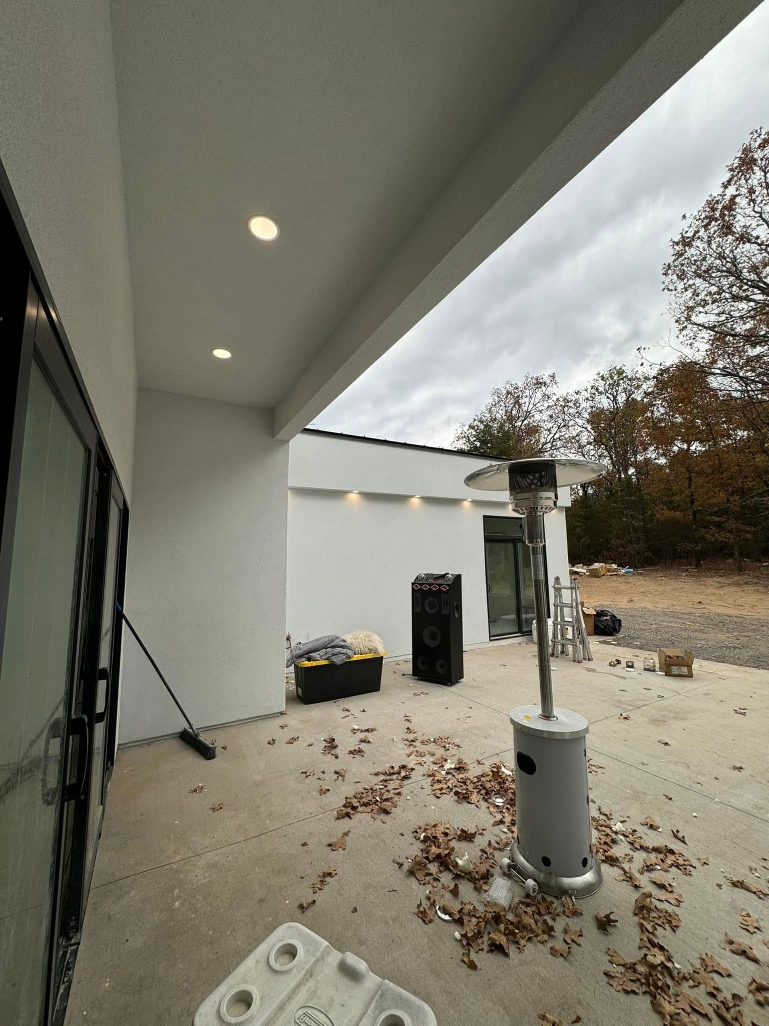 Outdoor patio with a heater, speaker, and construction materials. White walls, concrete floor, and cloudy sky.