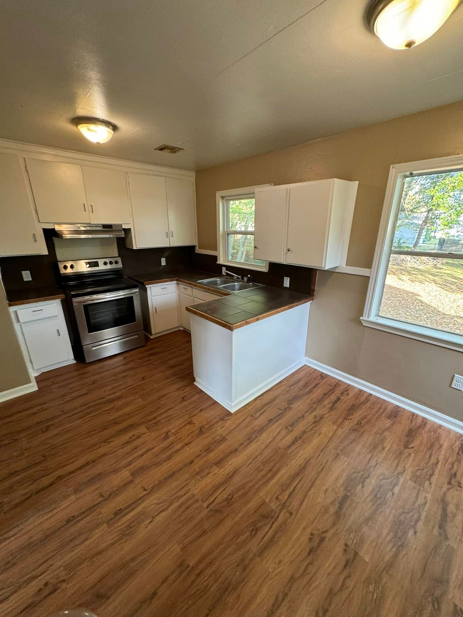 A kitchen with white cabinets, dark countertops, and wood-look flooring. Stainless steel oven, a small island, and two windows.