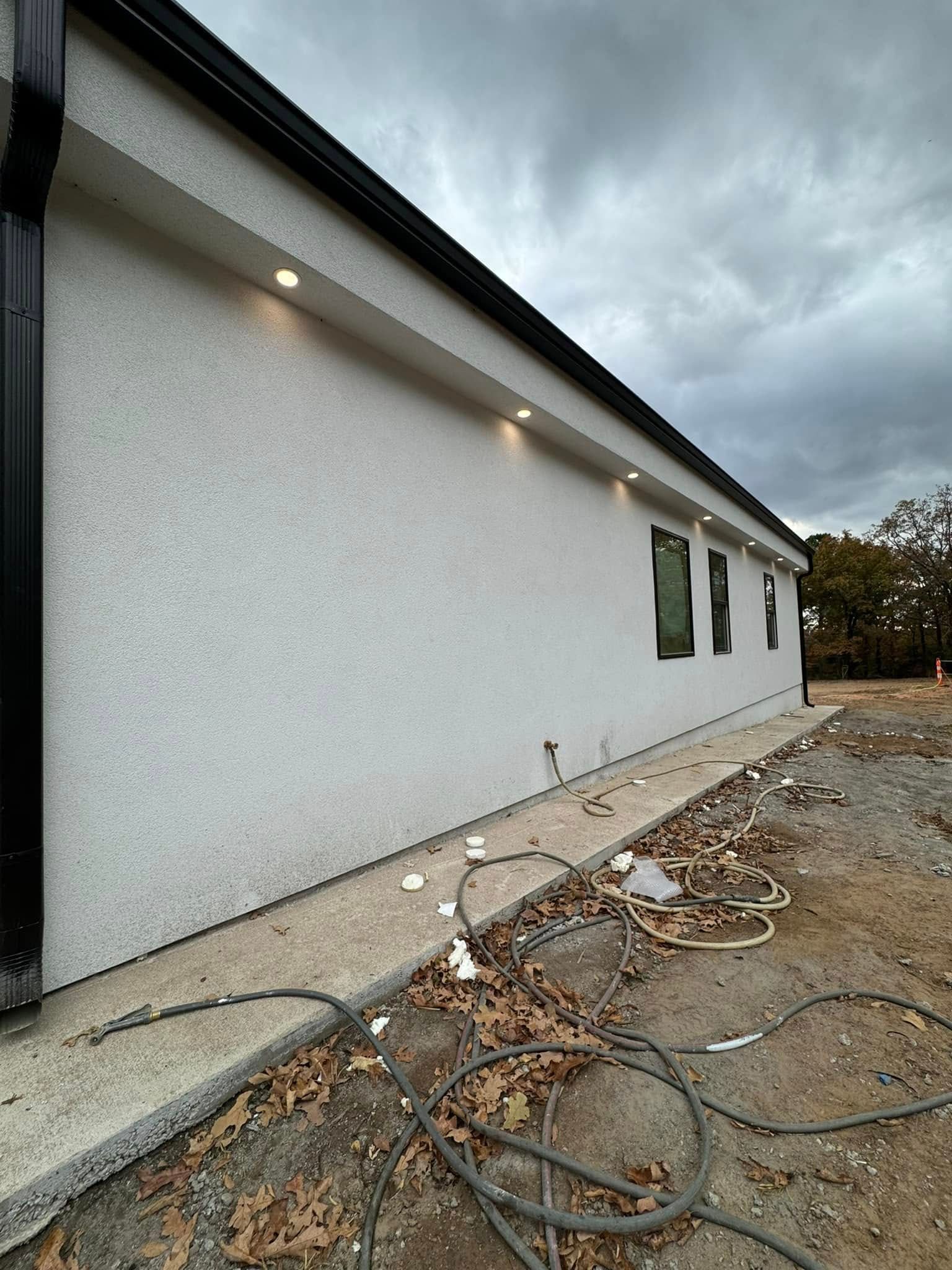Exterior of a building with white stucco walls, black trim, and recessed lighting. Construction debris surrounds the concrete base.