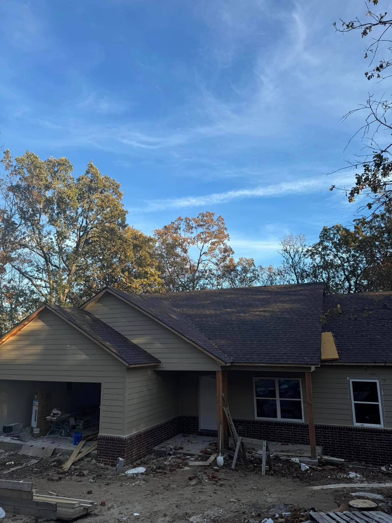 A house under construction with a dark gray roof and light green siding sits under a partly cloudy blue sky, surrounded by trees.