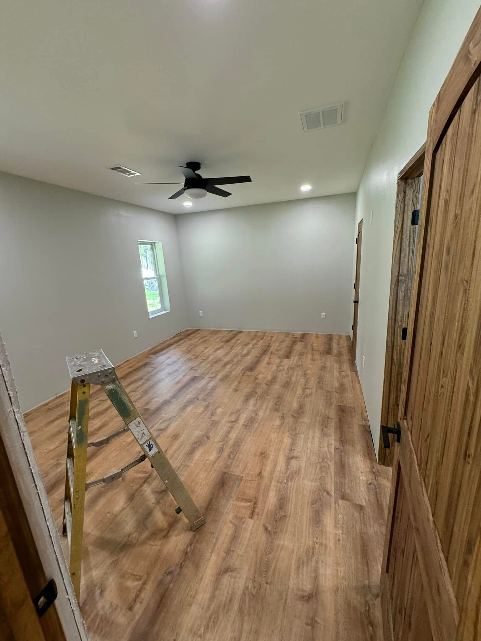 A room with wooden flooring, gray walls, and a ceiling fan. A wooden door and a ladder are present.