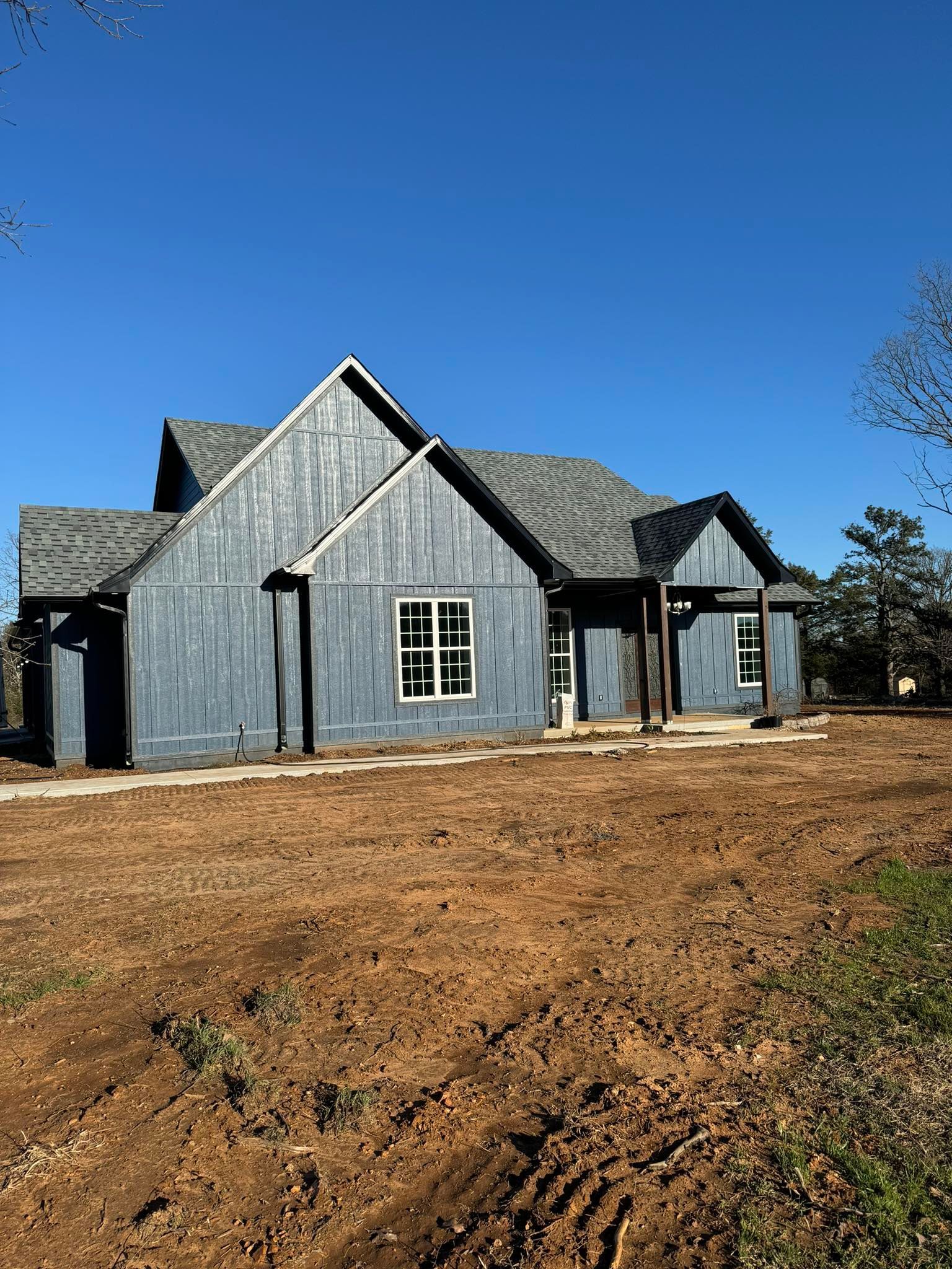 Newly constructed house with blue-gray siding under a clear blue sky. The yard is unlandscaped, with bare earth.